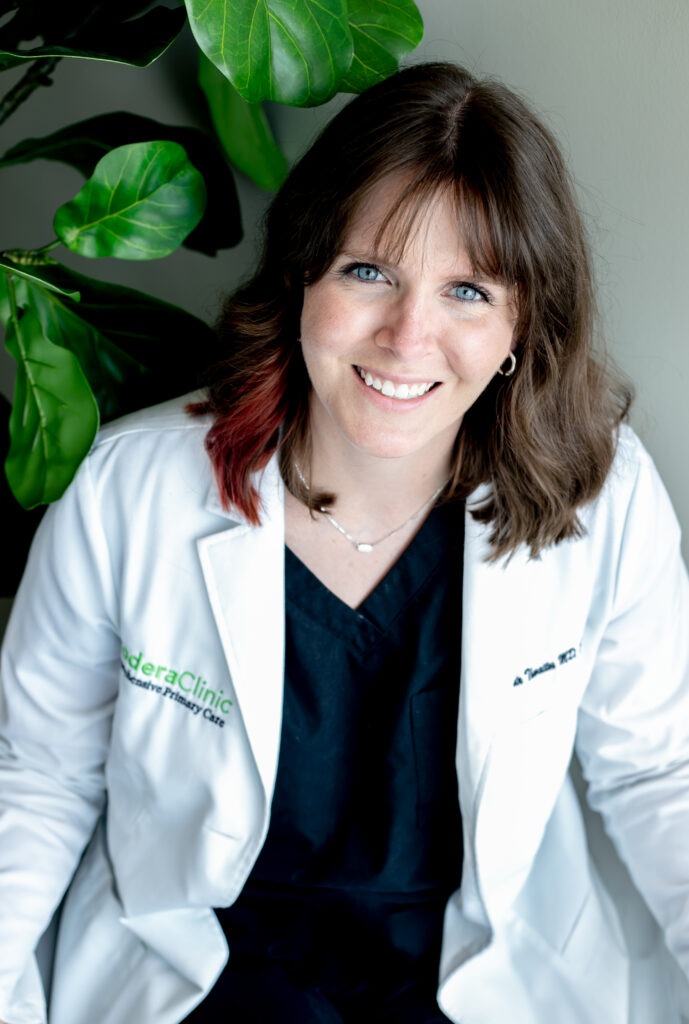 A woman with shoulder-length brown hair is smiling at the camera. She is wearing a white lab coat over a black outfit. There are green plant leaves in the background.