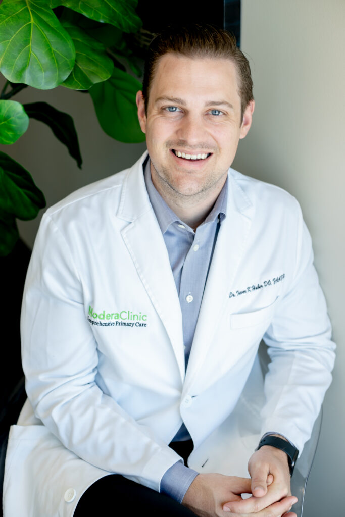 A doctor in a white coat, with "Modera Clinic" and "Integrative Primary Care" embroidered, smiles while seated. He wears a light blue shirt and watch. A large green plant is partially visible in the background.