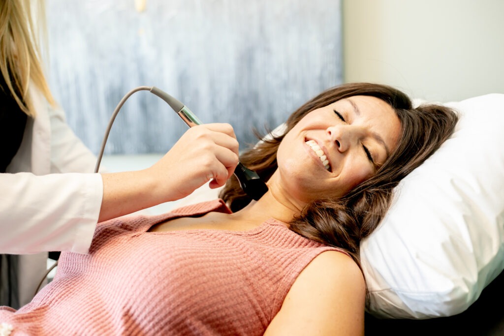 A woman is lying on a pillow, smiling, while a professional uses a handheld device on her neck. She appears relaxed in a medical or spa setting, wearing a pink sleeveless top.