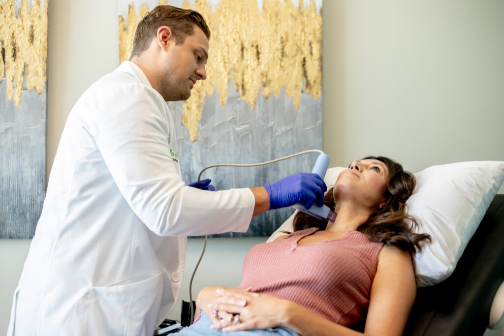 A healthcare professional in a white coat and blue gloves uses an ultrasound device on a woman's neck. The woman is lying on a bed with a relaxed expression. A large abstract painting is in the background.